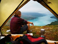 Hiking young man is sitting in front of a picturesque scenic view of lake Gjende and a remote mountain range in the Jotunheimen national park, Norway. Backpacker sitting in front of his tent gazing at stunning view. People traveling. XXXL (Sony Alpha 7R)